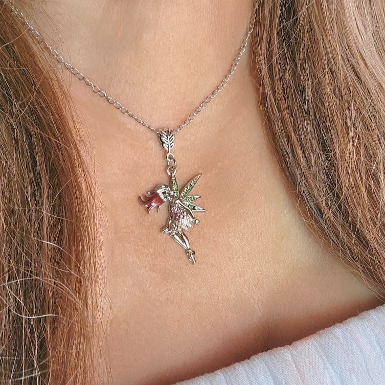 A fairy necklace with green and pink sparkles, worn around the neck. The fairy's red hair flows above a white top. The necklace sits perfectly against the skin, with light brown hair softly framing it.