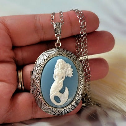 Held in hand, a mermaid cameo pendant necklace on a silver chain reveals exquisite detailing against a blue backdrop, invoking the elegance of oceanic tales.