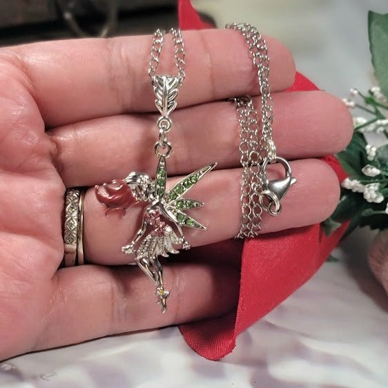 Hand holding a silver fairy necklace with shiny green wings and a pink dress. The fairy hangs from a silver chain next to a small heart charm. It's set against a white background with red ribbon and green leaves, giving it a romantic vibe.