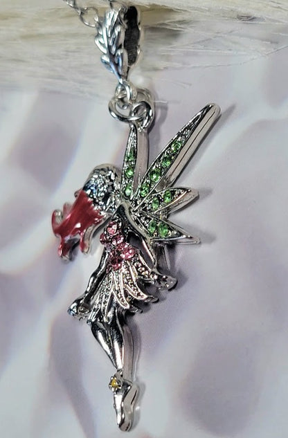 Silver fairy pendant with green glittery wings and pink crystal dress, featuring a touch of red on her flowing hair, attached to a shiny chain. The background is a soft, wavy white fabric that adds a magical touch.