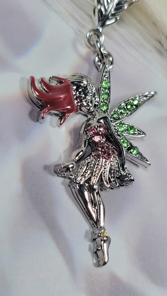 Silver fairy necklace charm with sparkly green wings, a red hair detail, and pink crystals on her dress. It's displayed on a light background that makes it shine.