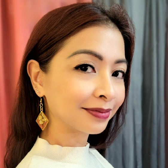 The image features a profile view of a woman with medium-length auburn hair wearing golden earrings, designed as a diamond-shaped piece encapsulating a vibrant yellow resin with botanical elements suspended within.