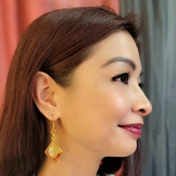 The image features a woman in profile view, showcasing an earring that is a radiant, translucent golden color with natural elements and glitter encapsulated within, hanging from a golden hook that adds a touch of sophistication.