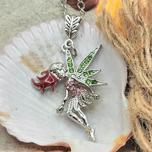 Silver fairy pendant with bright green wings and pink accents, lying on a seashell surrounded by sand, shells, and a piece of driftwood, bringing a touch of the ocean's magic.