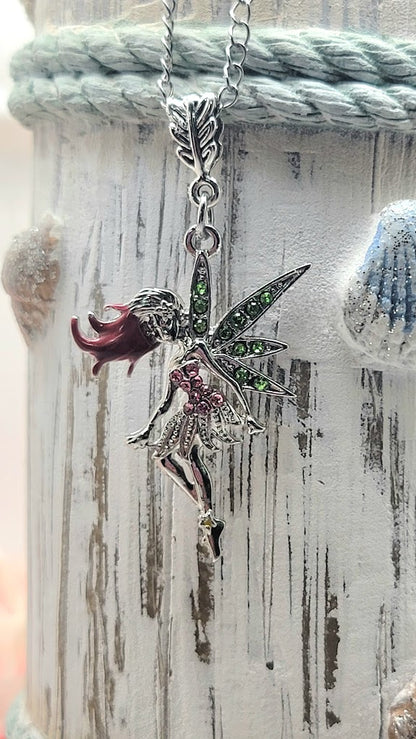 A glittery silver fairy necklace with green and pink details, hanging from a chain against a weathered wooden backdrop with a soft blue rope, giving it a vintage and whimsical feel.