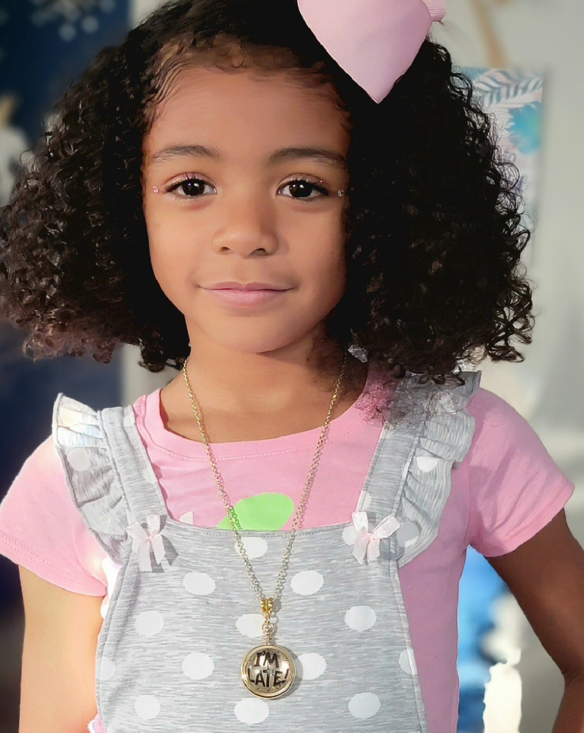 A bright-eyed young girl with bouncy curls and a pink bow wears a golden "I'M LATE" pendant necklace over a polka-dotted jumper, a combination of youthful innocence and whimsical style. 