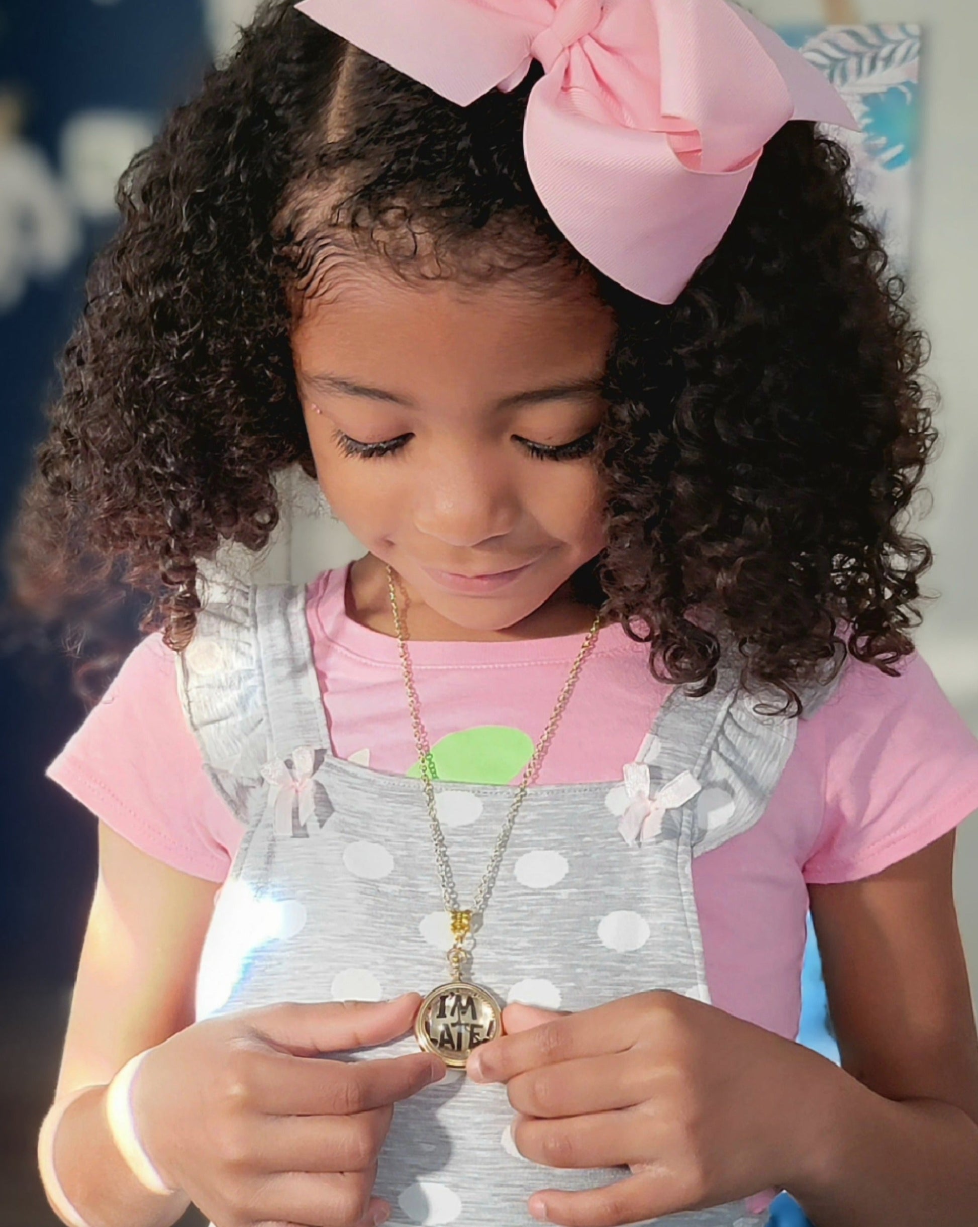 A young girl with curly hair and a pink bow looks at a golden "I'M LATE" pendant necklace she's wearing. The playful accessory contrasts with her polka-dot top, capturing a moment of childhood wonder inspired by a classic story.