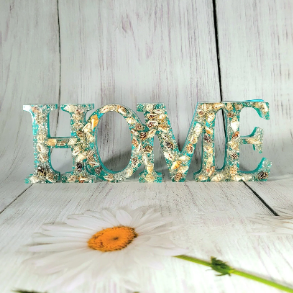 A captivating 'HOME' sign meticulously crafted with a blend of turquoise resin and genuine seashells encased within. Each letter reveals a myriad of seashells, both big and small, amidst a sparkling backdrop of glitter, evoking the serene vibes of the sea. The sign is set against a backdrop adorned with delicate flowers and botanicals, making it an artistic testament to coastal elegance.