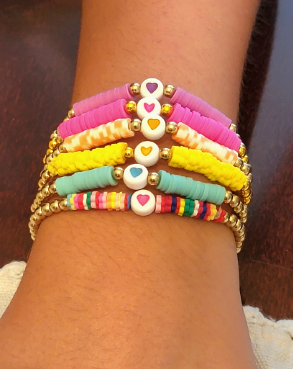 Stacked colorful beaded bracelets with heart charms, wrapped around a wrist against a wooden background.