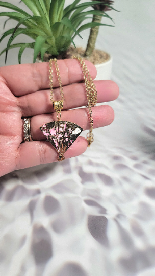 A hand gracefully holds a golden fan-shaped pendant with a cherry blossom design, its delicate chain glinting in the light against a natural backdrop.