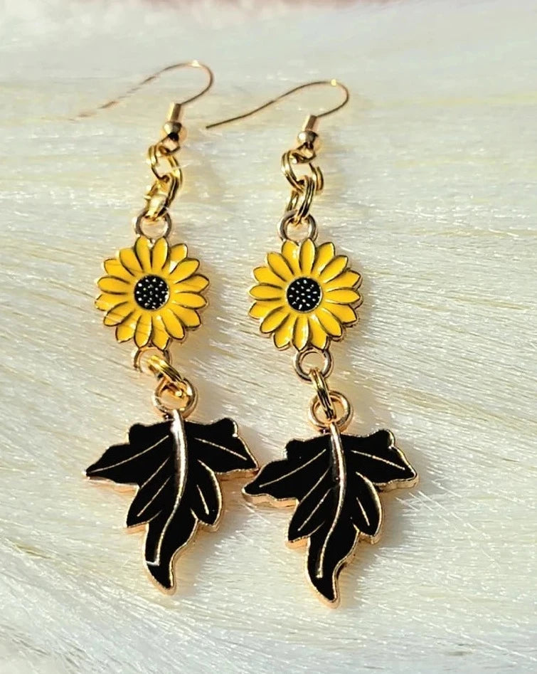 These earrings feature golden yellow sunflowers with intricate black centers attached to dark brown maple leaf charms. They exude a cheerful autumnal vibe against a softly textured background.