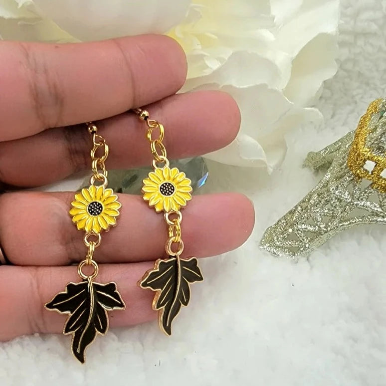 Golden earrings with a sunflower top and black leaves give a stunning twist to the autumn motif, held against a soft background, infusing a floral charm into the setting.