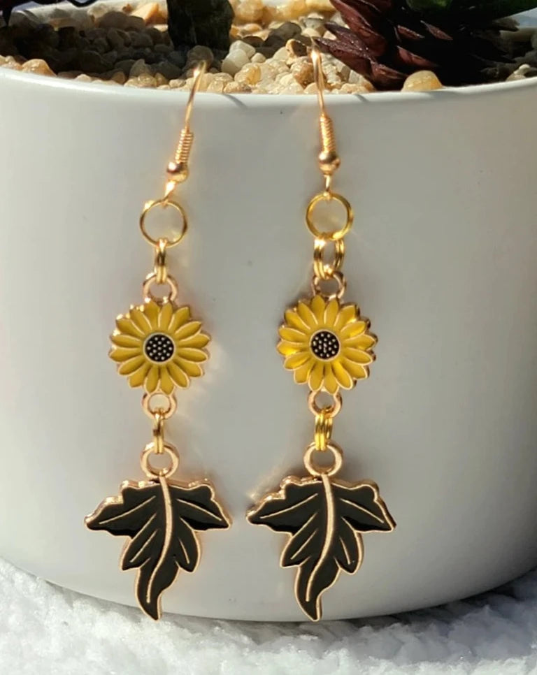 Sunlit golden earrings with cheerful sunflowers and elegant deep brown maple leaves dangle gracefully, set against a serene plant pot backdrop for a fresh, natural aesthetic.