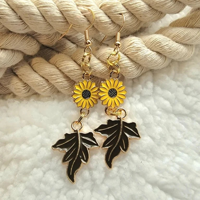 Vibrant golden sunflower earrings with dark brown maple leaves, bringing a burst of autumnal warmth, are set against a white braided rope and fluffy surface, marrying rustic texture with botanical beauty.