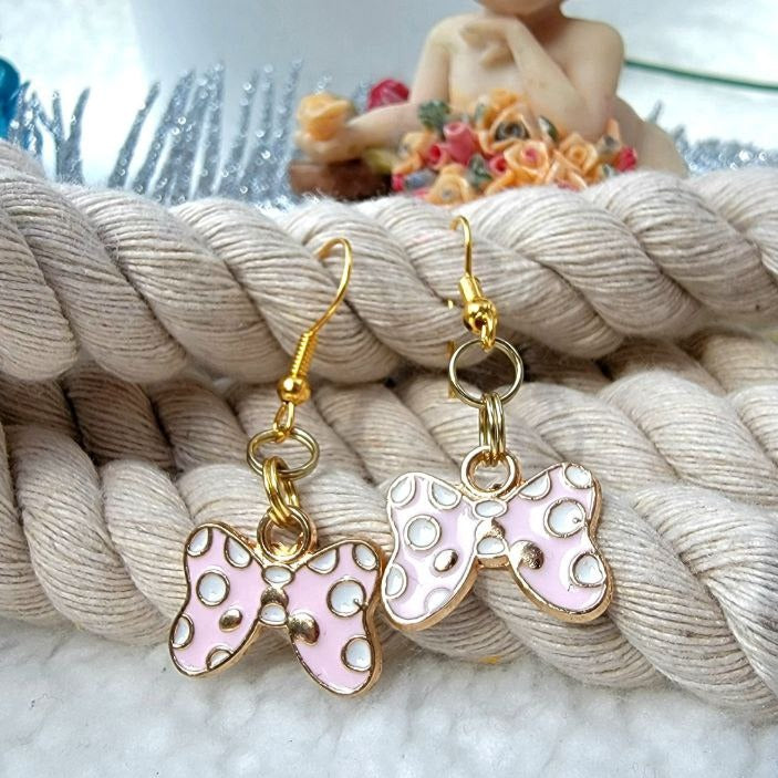 Whimsical golden bow-shaped earrings with pink and white enamel detailing, adorned with playful polka dots. They dangle gracefully against a braided backdrop, while a quaint figurine with florals nearby exudes an air of vintage charm.