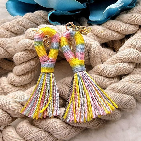 Multi-colored tassel earrings with shades of pink, blue, and yellow, presented on a soft white backdrop with green fabric leaves. The earrings feature gold hooks and are styled in a looped design.