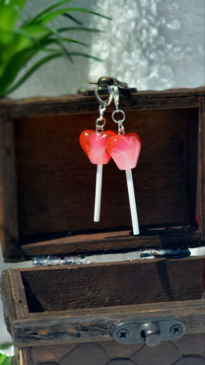 Vibrant red heart-shaped lollipop earrings with silver hoops, resting on a white textured background.