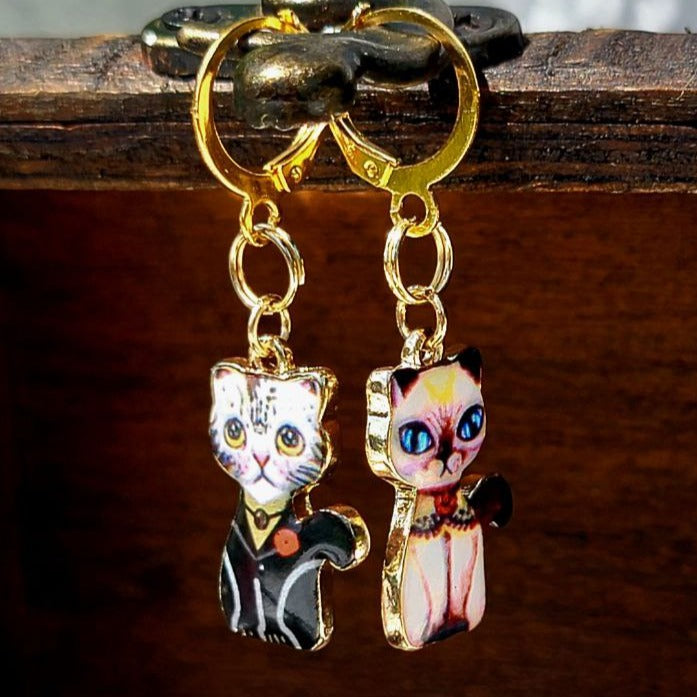 Elegant gold huggie earrings showcasing quirky cats: one with a black coat and heart patch, the other in pastel with striking blue eyes, both set on a fluffy white canvas.