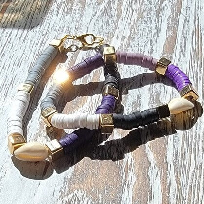 Two complementary bracelets lie side by side, one in tan and white and the other in black and purple heishi beads. Both are accented with small gold square beads for a touch of harmony and subtle elegance. 