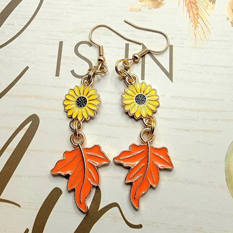 Sunflower earrings with leaf charms evoke the cheerful spirit of autumn, their golden and orange hues standing out against a script-patterned surface.