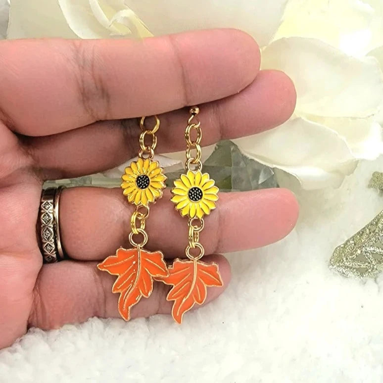 A hand cradles a pair of charming earrings featuring golden sunflowers connected to vibrant orange leaves, all against a soft, fluffy white backdrop.