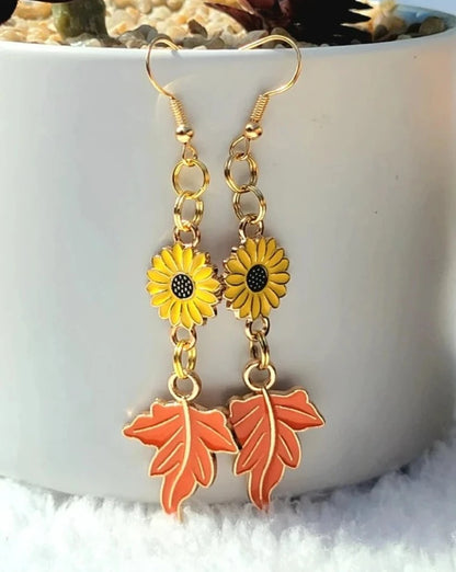 Golden sunflower earrings with orange leaf pendants dangle against a snowy fabric, with soft petals and a lush plant providing a vivid backdrop.