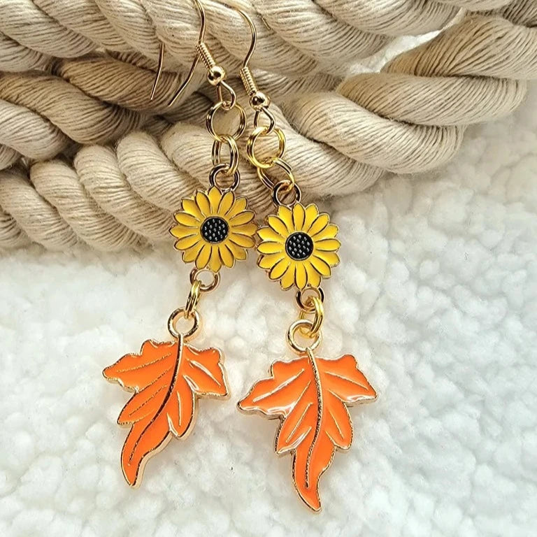 These charming earrings capture the essence of autumn, featuring golden yellow sunflowers linked to vibrant orange maple leaves, dangling from golden hooks and resting against a nautical rope background for a rustic touch.