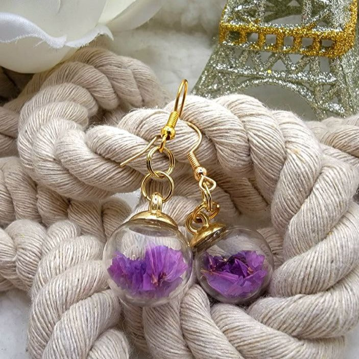 Delicate gold earrings featuring clear orbs encapsulating vibrant purple flowers, draped over a cream woven rope. In the distance, a soft white flower and a glittering Eiffel Tower ornament enhance the romantic ambiance.