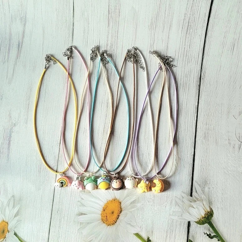 A collection of pastel corded necklaces with whimsical charms lay on a rustic white wooden backdrop. Each necklace showcases delightful motifs like rainbows, smiling stars, and cute animals. The serene display is enhanced by soft daisy blooms.