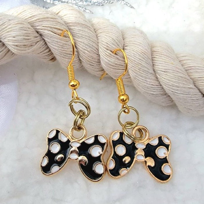 Black enamel bow-shaped earrings with white polka dots and gold trim, hanging gracefully against a beige braided texture with glimmering silver tassels above.