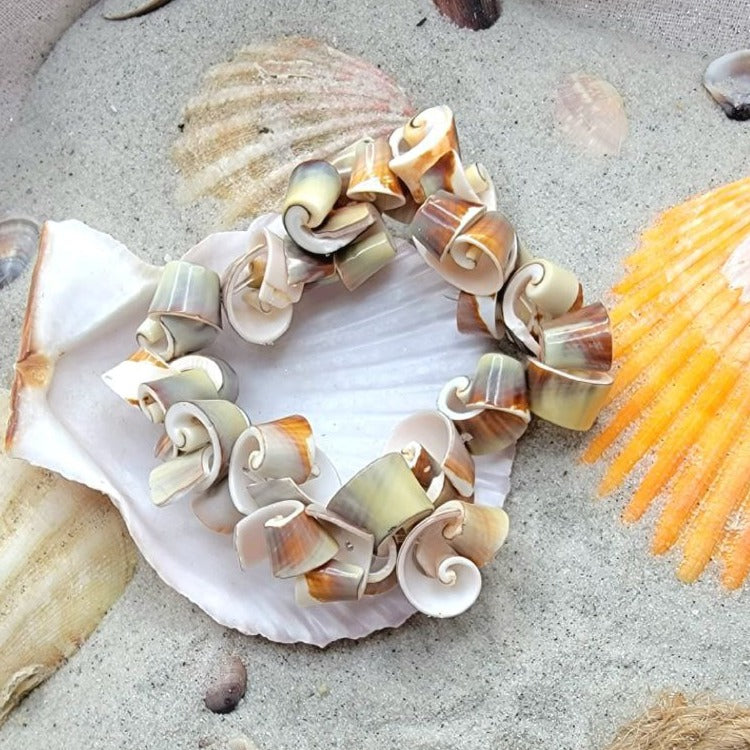 An intricately crafted bracelet made of natural spiral seashells, each uniquely shaded with tones of cream, brown, and soft yellow. It's laid on a sandy background adorned with an array of larger seashells and rustic marine elements.