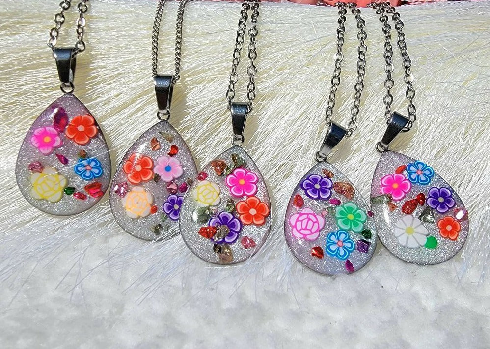 A collection of vibrant teardrop-shaped pendants, each adorned w/ colorful flowers set against a glittery backdrop, dangle from dark chains. They lay atop a soft-textured surface, inviting a sense of whimsical elegance.