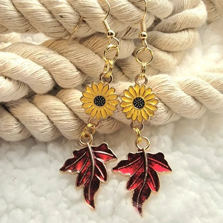 Golden sunflower charms paired with deep red autumn leaf pendants make a charming statement on these earrings, highlighted by a soft, textured background.