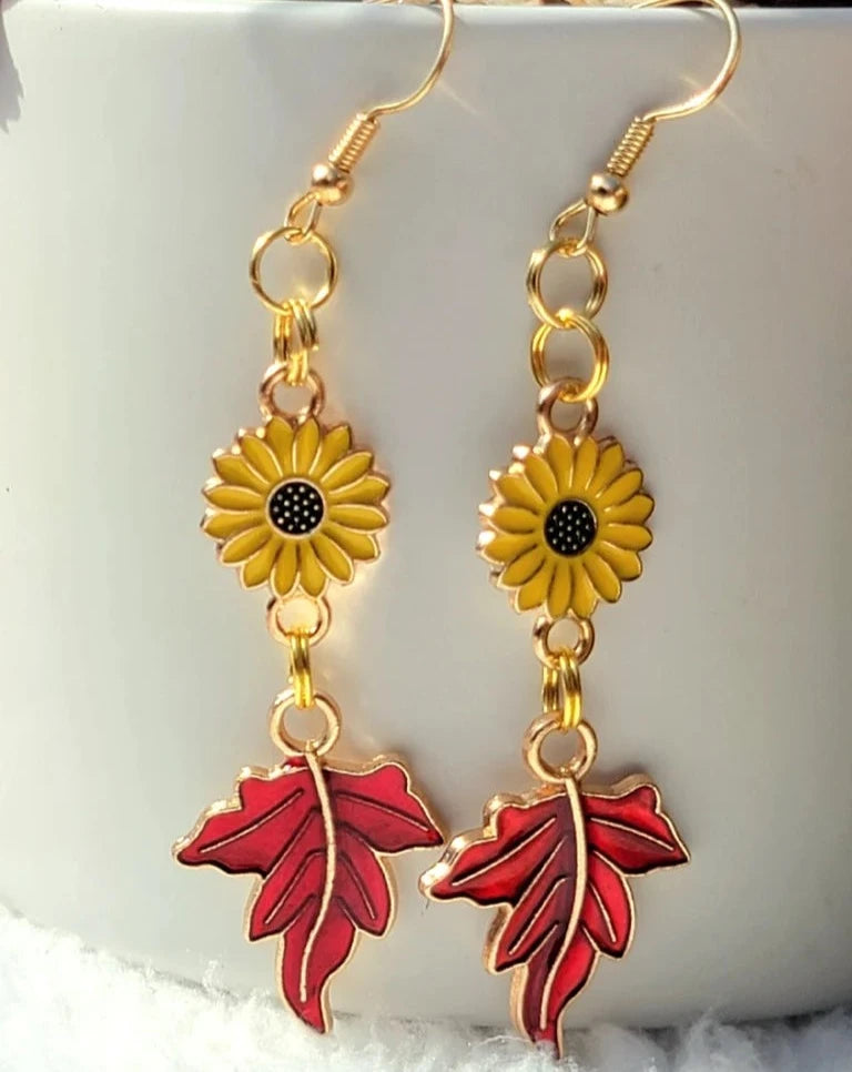 Bright sunflower earrings with vibrant red maple leaf pendants are held up, capturing the essence of fall's warmth against a soft backdrop of snow.