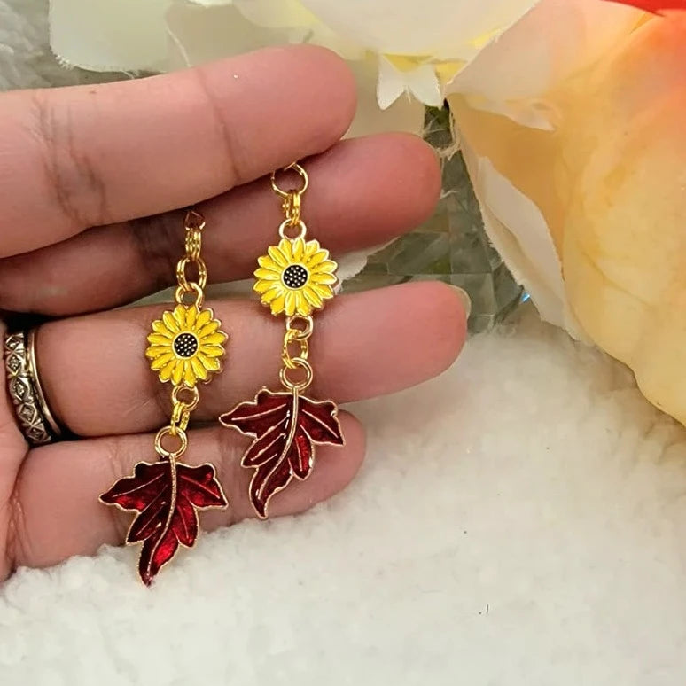 Golden sunflower earrings with red autumn leaf charms are gracefully presented on a hand over a soft bed of white fluff, evoking a cheerful fall day. 