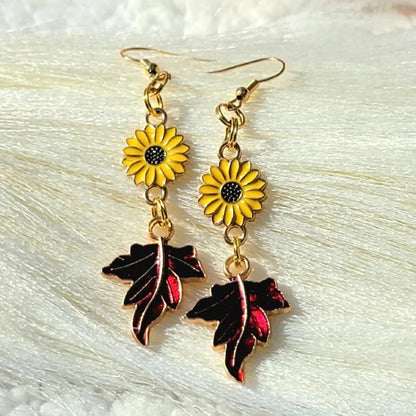 Vibrant yellow sunflower charms with black centers dangle above glossy red autumn leaves on these earrings, set against a snowy white backdrop. 