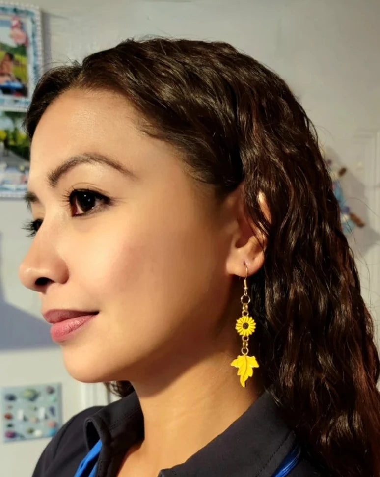 A model displays gold dangle earrings, each adorned with a sunflower and autumn leaf charm. The elegant jewelry, set against a wintry backdrop, evokes the transition from summer's warmth to fall's crispness.