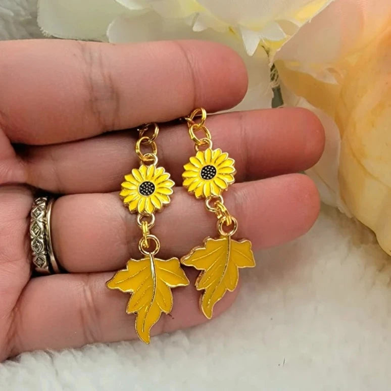 Golden sunflower charms with black centers dangle above textured maple leaf pendants on these earrings. Held between fingers, they exude autumn's warmth against a white, fluffy background. 