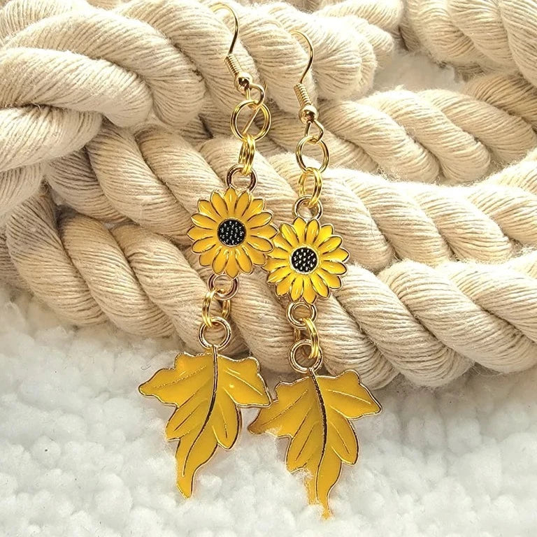 Vibrant sunflower earrings with intricate black centers linked to delicate golden maple leaves presented on a soft, twisted cream rope. A cheerful homage to fall's lively palette. 