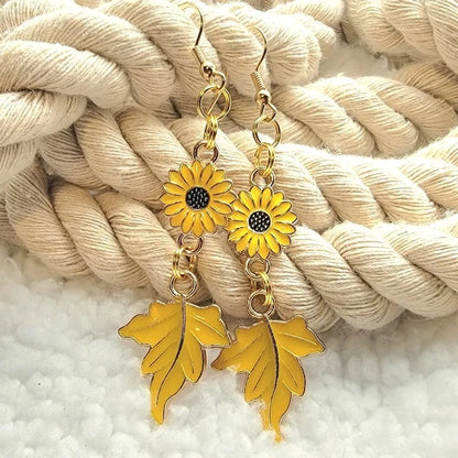 Vibrant sunflower earrings with intricate black centers linked to delicate golden maple leaves presented on a soft, twisted cream rope. A cheerful homage to fall's lively palette. 