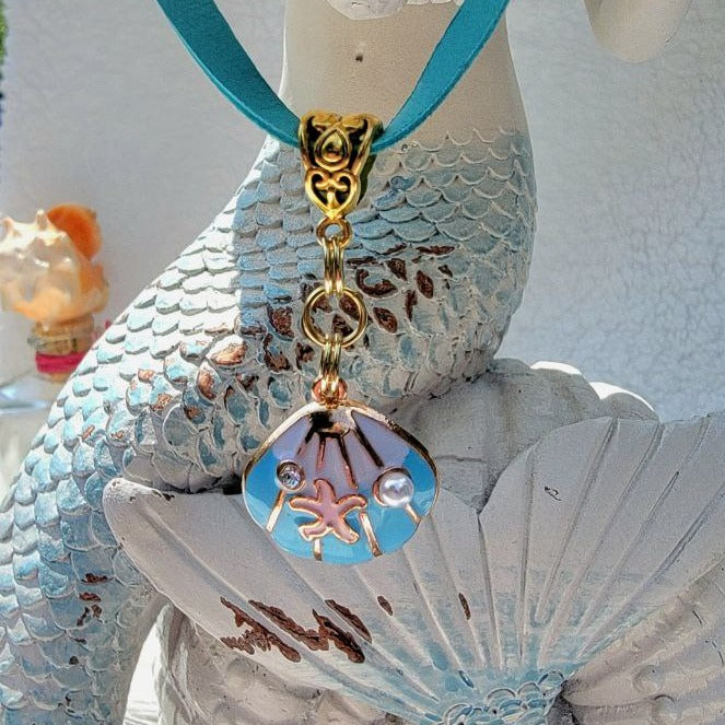 A vibrant turquoise-colored leather cord showcases a delicate clam or blue oyster charm with a central starfish motif. The charm dangles gracefully from a detailed Tibetan bail and is displayed against a textured mermaid statue with intricate scales and rustic white and blue patina.